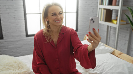 Woman smiling in red pajamas taking selfie in modern bedroom with bright window and cozy bed...
