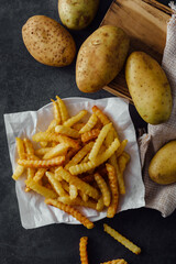 Fresh Crinkle Cut French Fries Served with Raw Potatoes on Dark Surface