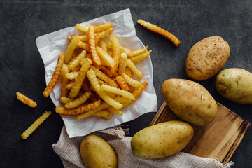 Crispy French Fries on White Paper with Raw Potatoes on Dark Stone Surface