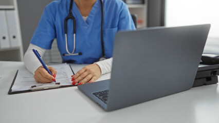 Woman in blue scrubs writing in a hospital room, hands on a clipboard with a laptop nearby, suggesting a professional medical setting for adults.