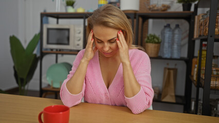 Woman stressed at workplace with short blonde hair in an office room, wearing pink sweater,...