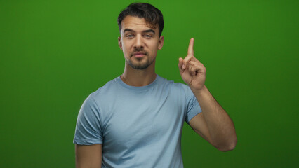 Fototapeta premium Young hispanic man points upward with closed eyes wearing blue shirt against vibrant green background suggesting an idea or thought.