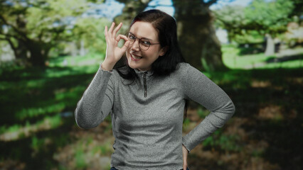 Woman with glasses in park flashes playful hand sign in bright outdoor setting surrounded by green...