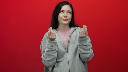 Woman smiling in gray hoodie making money gesture against red background showing playful confidence...