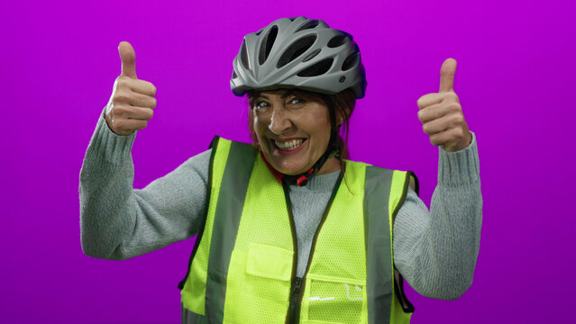 Woman smiling with thumbs up wearing helmet and safety vest against pink background