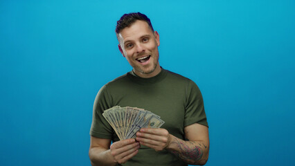 Young man holding american dollars against a vivid blue background appears joyful and content,...