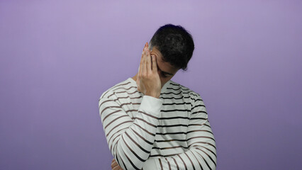 Young man in striped shirt against a purple background looks stressed with hand on face suggesting...