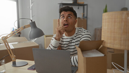 Young man in a striped shirt sitting at a desk in a new home office surrounded by boxes, looking thoughtful while using a laptop, lamp casting light on the workspace.