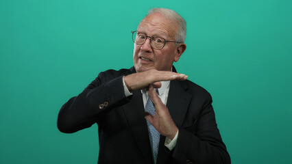 Senior man in a business suit makes a timeout gesture against a vibrant green background, conveying...