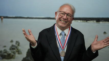 Senior man in a suit smiling with a medal at a seaside beach, celebrating success against a background of water and rocks.