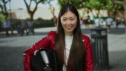 Woman with long hair in red jacket holds helmet while standing in urban street with trees and...
