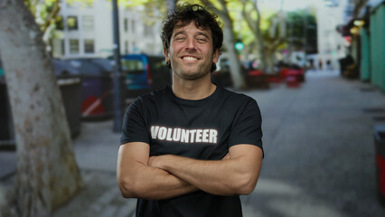 Hispanic man with a handsome smile and folded arms wearing a volunteer shirt standing confidently...