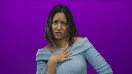 Obraz premium Young woman with hand on chest appears surprised while standing against a vibrant purple background, wearing a light blue sweater, conveying a sense of disbelief and emotion