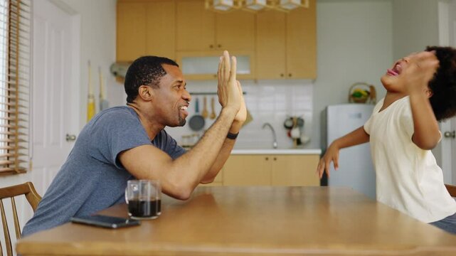 Cheerful Black African American father and little child boy smiling and sharing a joyful high five across a dining table in kitchen, happiness and connection through playful interaction