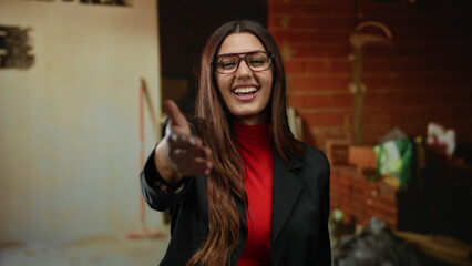 Hispanic woman in glasses smiling and extending hand inside construction site, wearing red shirt...