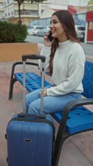 Woman sitting on bench talking on phone with suitcase nearby in urban street setting showcasing casual travel and hispanic style.