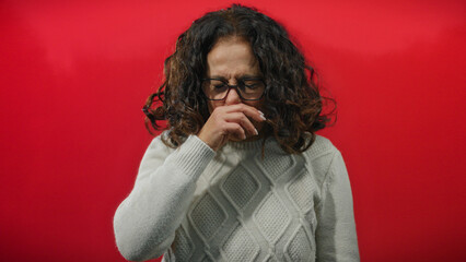 Hispanic woman with curly hair and glasses sneezing over isolated red background wearing a white...
