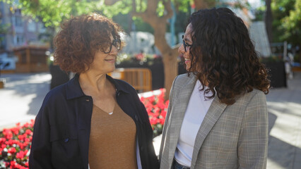 Women talking in a city park with bright flowers and trees, reflecting friendship and happiness in an urban outdoor setting on a sunny day.