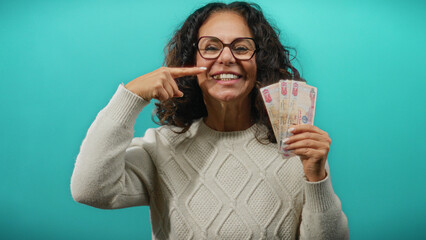 Middle-aged woman in glasses holding uae dirhams against blue wall, smiling and pointing at her...