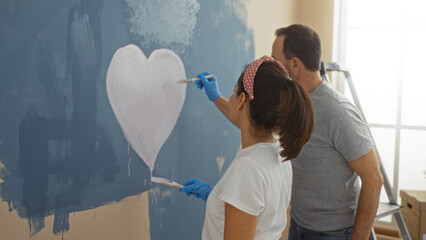 Middle-aged hispanic couple painting a heart on a wall together in their cozy new home living room, showcasing love and teamwork in an indoor setting.