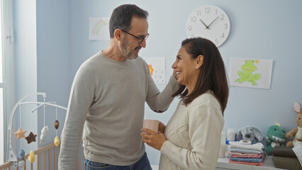 Obraz premium Middle-aged hispanic couple in a cozy bedroom shares a loving moment by a cradle, with the woman holding a coffee cup and children's drawings adorning the walls.