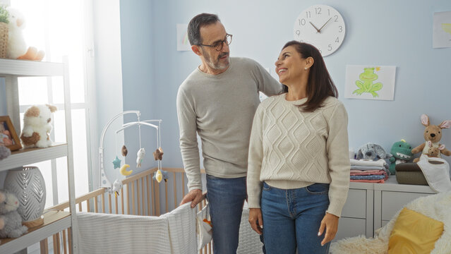 Hispanic middle-aged couple smiling in a cozy baby room with a cradle, surrounded by toys and soft lighting, showing love and family warmth indoors.