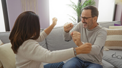 Middle aged hispanic couple laughing joyfully indoors in a cozy living room setting, conveying love and happiness.