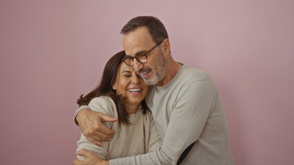 Middle-aged hispanic couple embraces lovingly against a pink wall, wearing casual attire,...