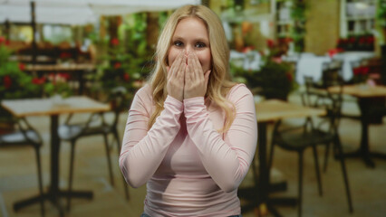 Fototapeta premium Young blonde woman in pink top playfully covers her face and smiles at a restaurant with outdoor tables and vibrant foliage.