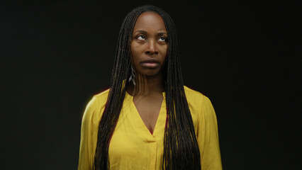 African woman wearing yellow shirt stands against a dark background, showcasing her braided...