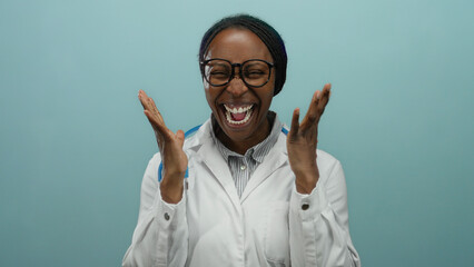 Smiling african woman doctor in white coat with stethoscope celebrates joyfully against blue wall...
