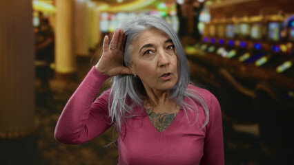 Woman in casino with grey hair gesturing listening in front of slot machines, conveying curiosity and contemplation in a lively gambling environment.