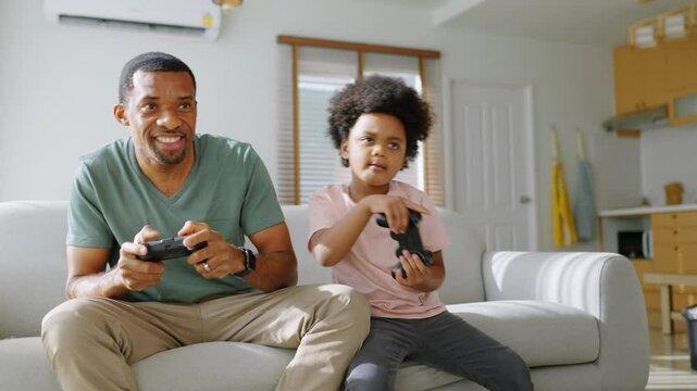 Cheerful Black African American Father and his Little son in Afro hair sitting on sofa using joysticks or game controllers playing console video games at home - Powered by Adobe