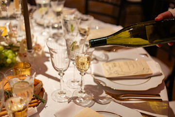White wine is gracefully poured into a crystal glass at a beautifully arranged dinner table, featuring fine china, menus, candles, and golden-hued celebratory ambiance