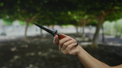 Male hand holding a knife in a park setting with blurred trees in the background, depicting an...