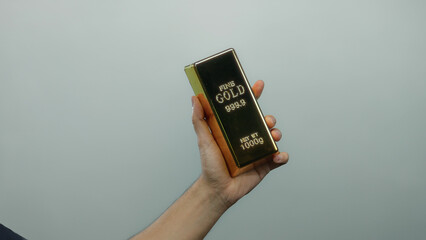 Man holding a shiny gold bar in front of a plain white background, emphasizing wealth and...