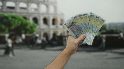 Male hand holding romanian banknotes near the roman coliseum, capturing a fusion of culture,...