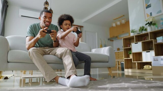 African Father and son with excited expressions on a couch playing a video game with controllers in hand, showing joyful and competitive expressions in living room.