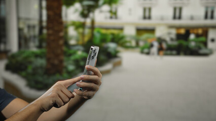 Man using smartphone on city street outdoors with greenery and buildings in the background showcasing urban lifestyle and technology interaction.