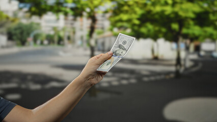 Man holding american dollar banknote in city street under sunlit trees, highlighting concepts of...