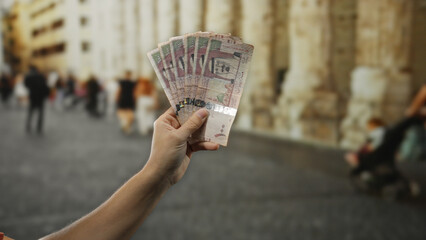 Man holding saudi arabian riyal banknotes outdoors on busy city street, showcasing currency's...