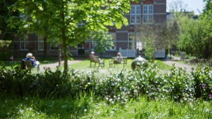 Obraz premium Blurred park scene with people seated on benches surrounded by lush greenery and a historic building backdrop, creating a peaceful, relaxed atmosphere with a touch of nature.