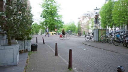 Blurred view of a european city street with a woman biking in the foreground and a historic townscape in the background surrounded by lush green trees on a sunny day.