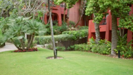 Blurred view of a lush outdoor garden scene with vibrant greenery and red buildings in a tropical resort, featuring a defocused luxurious background setting under sunny skies.