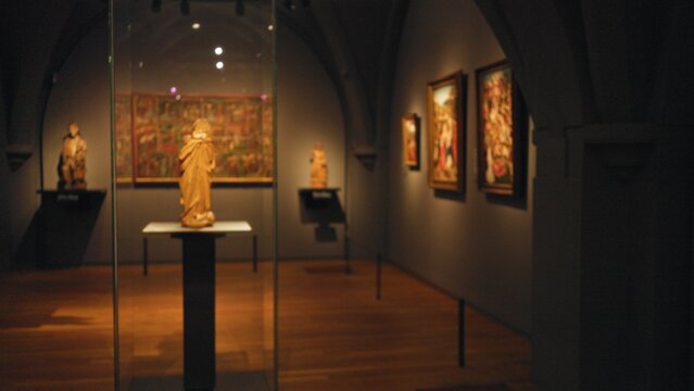 Blurred museum scene indoors with focus on sculptures and paintings surrounded by dim lighting and wooden floor, creating an artistic and contemplative ambiance.