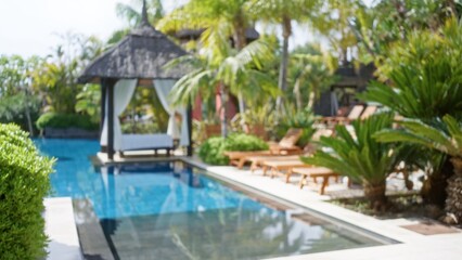 Blurred image of a tropical resort with palm trees, a luxury pool, sun loungers, and a shaded cabana exhibiting a tranquil ambiance under clear skies.
