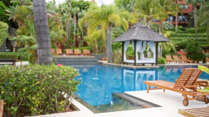 Defocused view of a luxury resort showcasing a tranquil poolside setting with palm trees, wooden lounge chairs, and a serene tropical background under a clear blue sky.