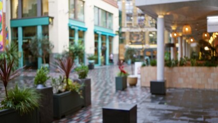 Defocused view of a london street with plants and shops in winter, capturing a blurred, urban scene with outdoor seating and wet pavement under soft bokeh lighting.