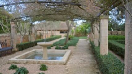 Blurred garden scene featuring a mediterranean style, defocused fountain and pathway under bokeh light, creating a serene atmosphere with lush greenery.