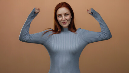 Woman with red hair smiling confidently and flexing arms in blue sweater against a brown wall...